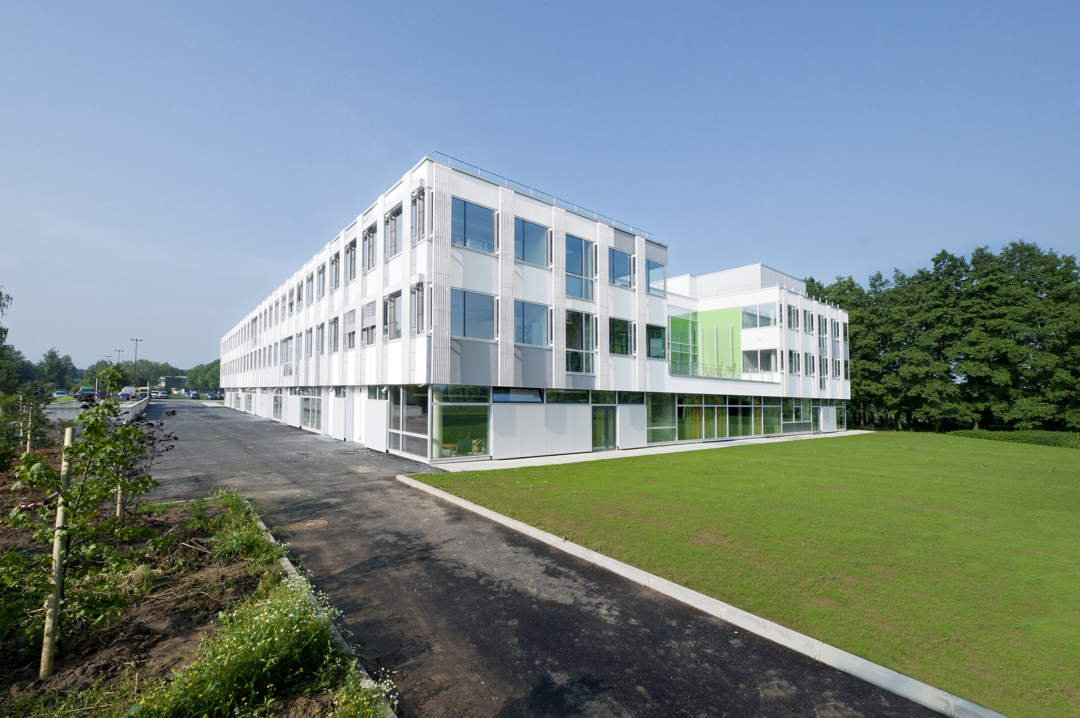 The white modern building with large windows and green accent facade, set beside a lawn and paved path under a clear blue sky.