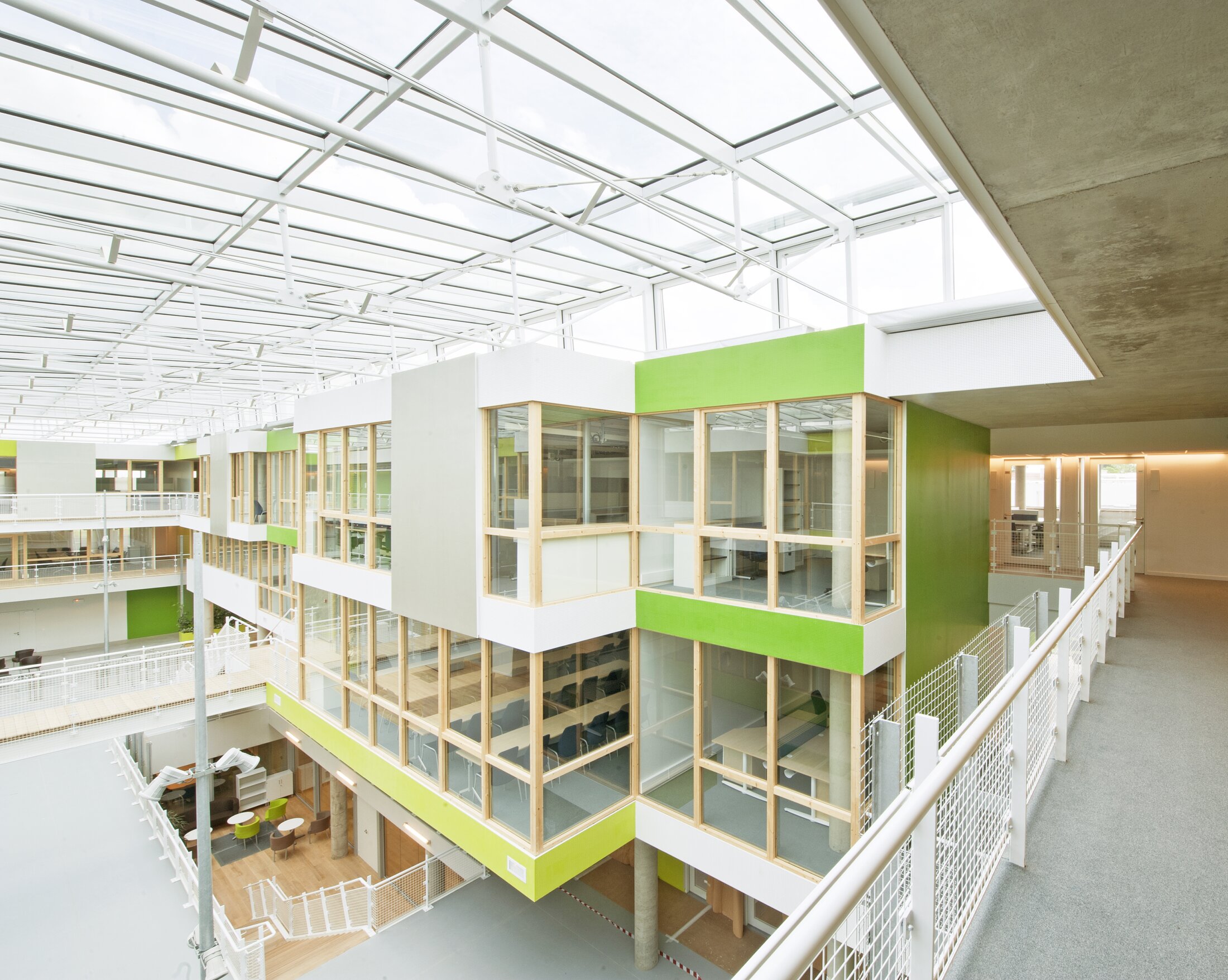 Project by Behnisch Architekturbuero, Digiteo Labs - 3 Research Buildings. Bright atrium with glass roof and green-accented offices, connected by walkways and railings around an open central space.