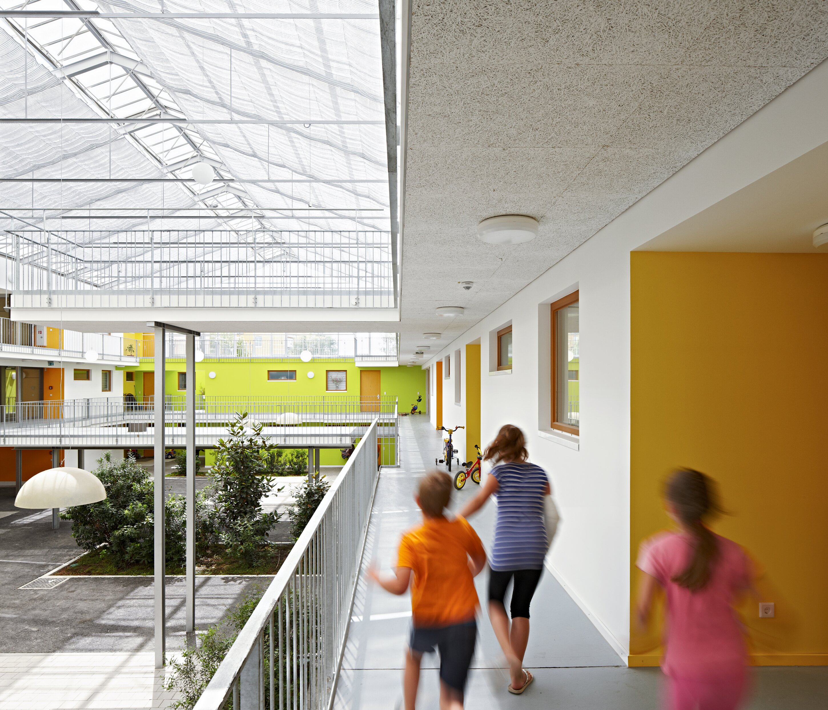 Project by Behnisch Architekturbuero, Residential Building Ingolstadt-Hollerstauden. Children run along the open corridor overlooking the shared courtyard in the building with colorful walls and a glass roof.