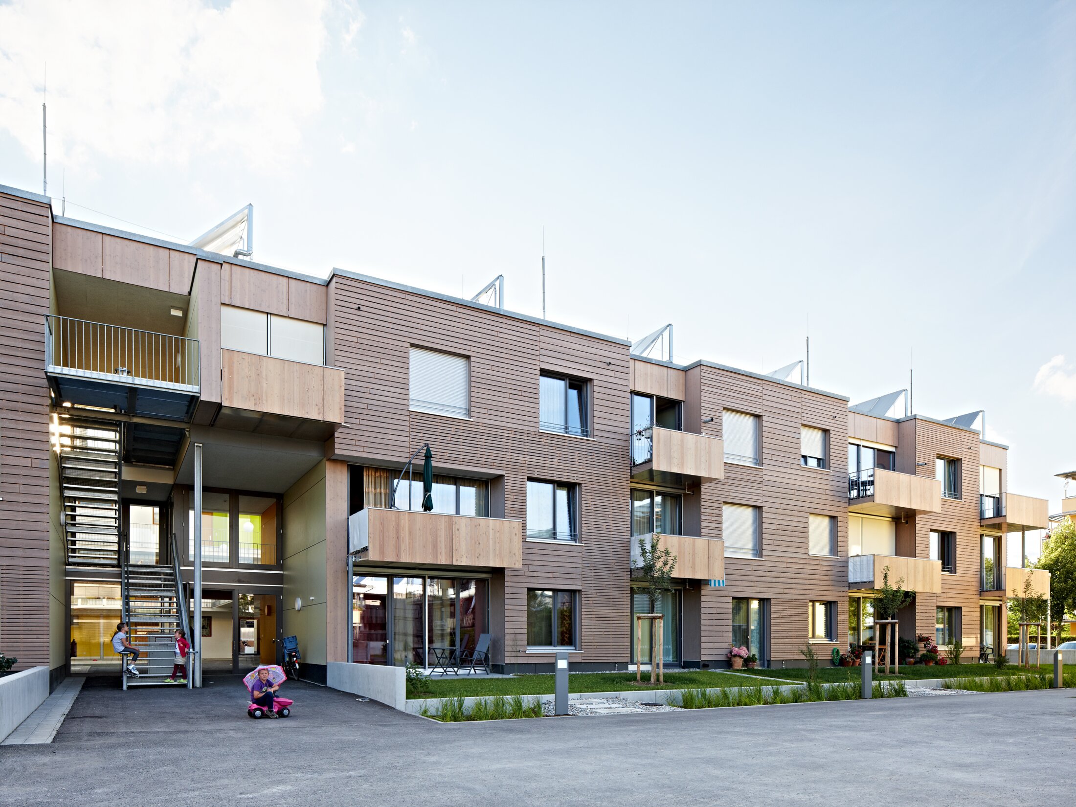 Project by Behnisch Architekturbuero, Residential Building Ingolstadt-Hollerstauden. Modern apartment building with wood facade and balconies, small courtyard, and children playing near an exterior stair.