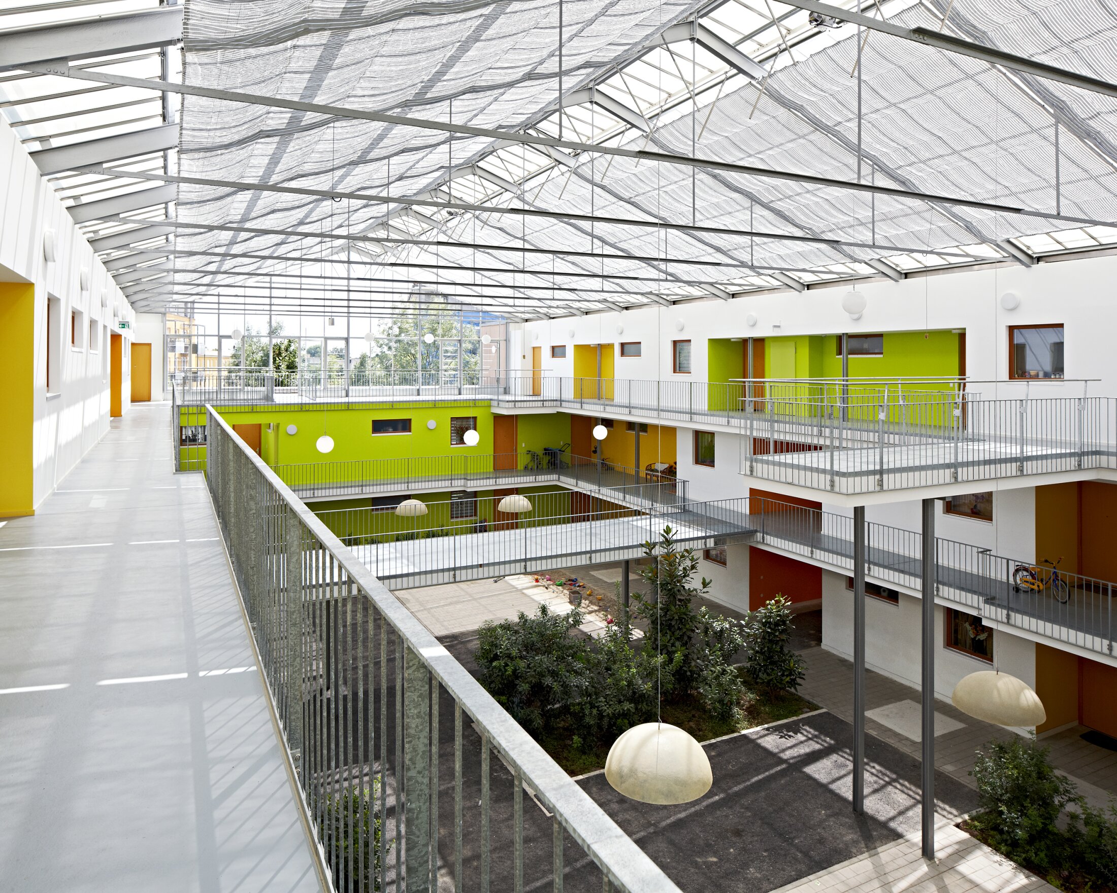 Project by Behnisch Architekturbuero, Residential Building Ingolstadt-Hollerstauden. Interior courtyard of the building with open walkways, green, orange and yellow walls, railings, and a glass roof letting in daylight.