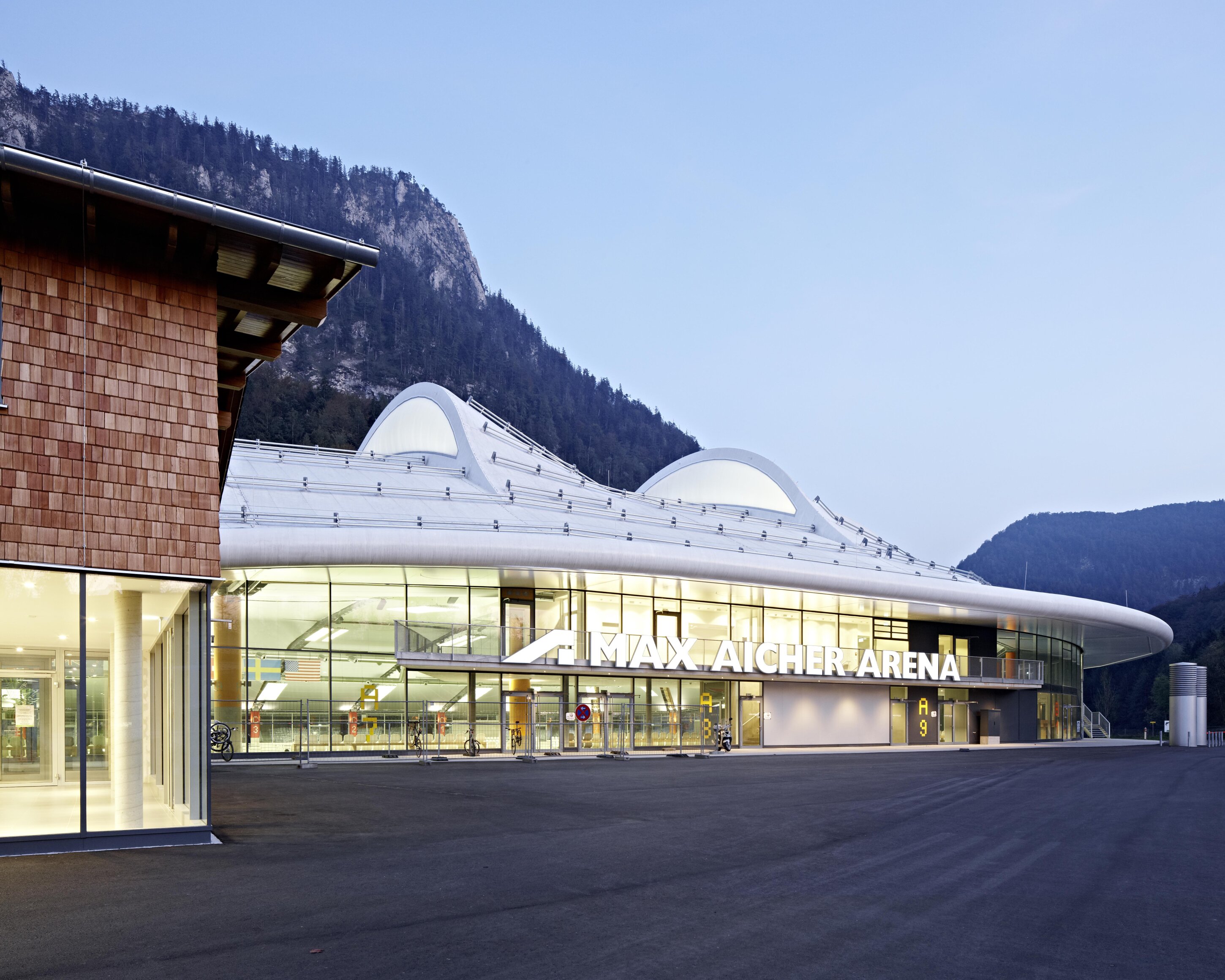 Project by Behnisch Architekturbuero, Max Aicher Arena. Modern arena building with curved roof and glass facade, labeled &ldquo;Max Aicher Arena,&rdquo; set against mountains at dusk.