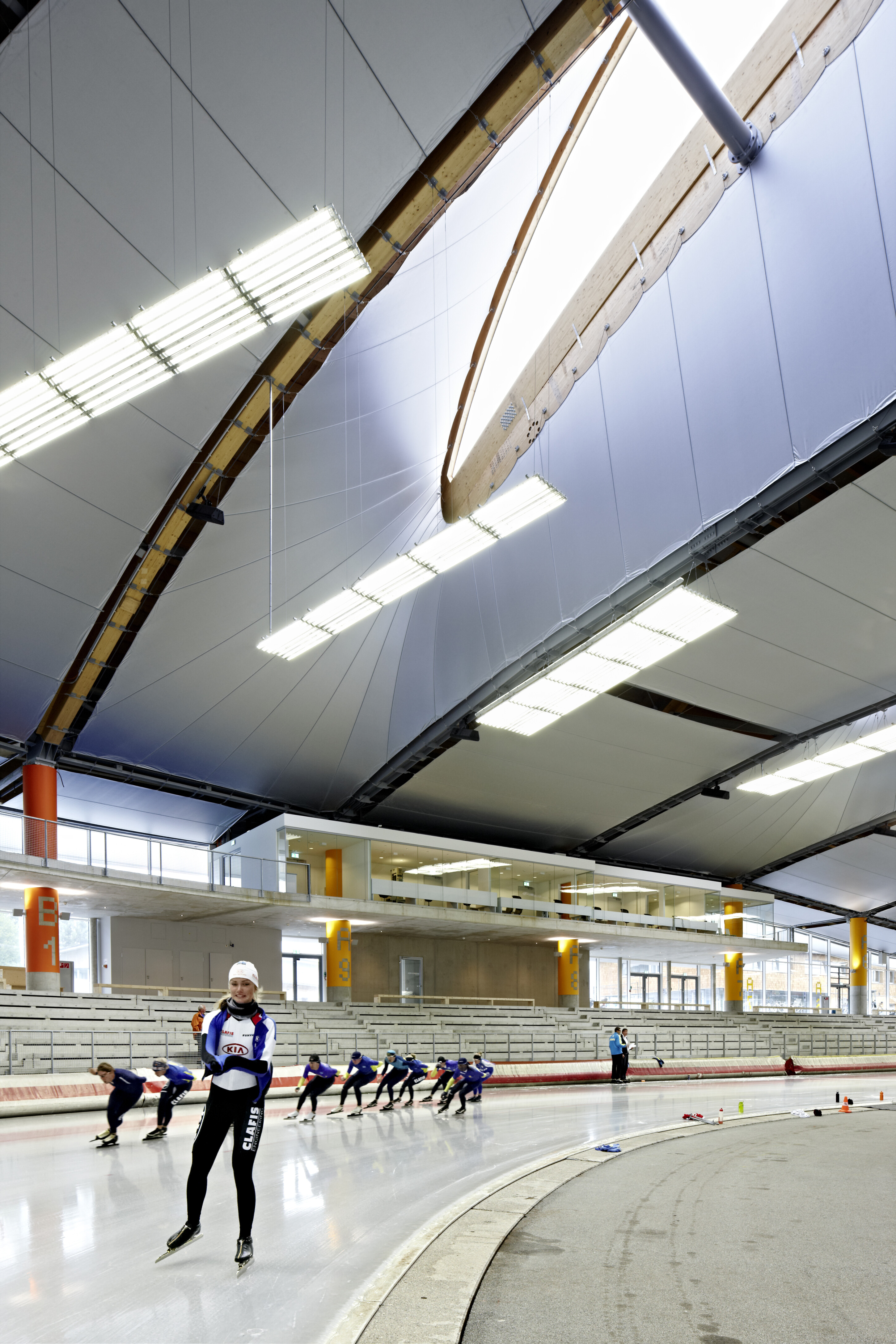 Project by Behnisch Architekturbuero, Max Aicher Arena. Speed skaters are training in an ice rink. One skater is visible in the foreground, the others are skating a curve. The roof with a large skylight is visible at the top of the picture.