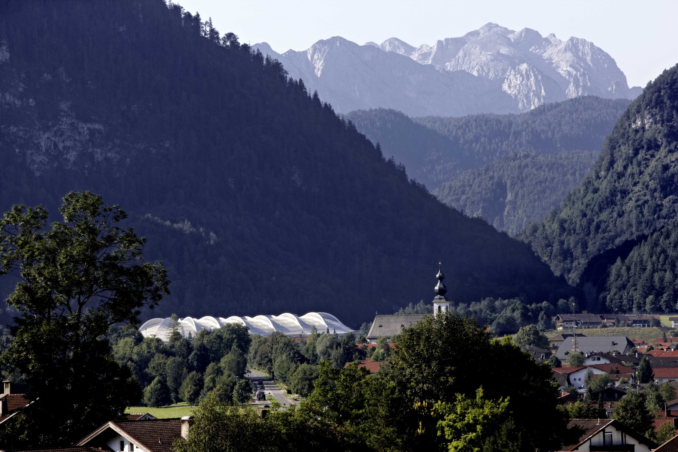 Project by Behnisch Architekturbuero, Max Aicher Arena. Mountain valley with forested slopes, a village of houses and a church steeple, the white-roofed building in the distance.