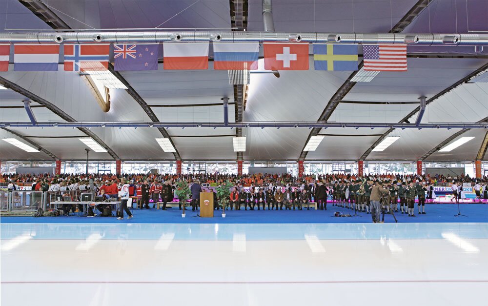 Project by Behnisch Architekturbuero, Max Aicher Arena. Ceremony on the indoor ice rink with a large crowd and musicians on the floor. International flags are hanging overhead.