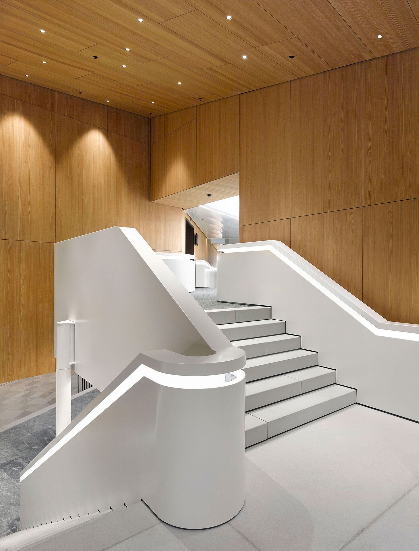 Project by Behnisch Architekturbuero, WIPO Conference Hall. Interior stair with white curved railings and steps, surrounded by wood-paneled walls and ceiling with recessed lighting.