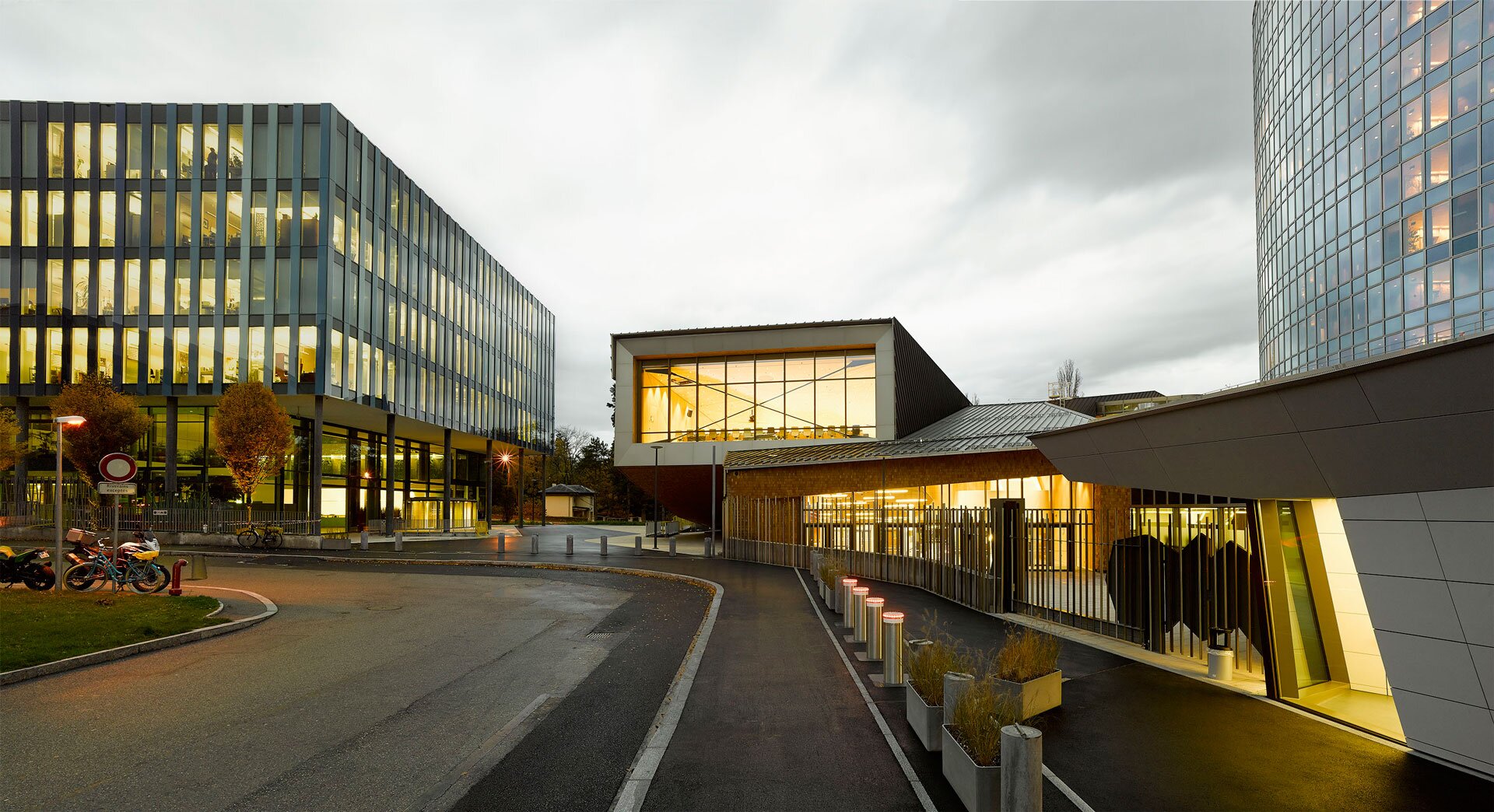 Project by Behnisch Architekturbuero, WIPO Conference Hall. Modern office buildings at dusk with illuminated interiors, a curved road in the foreground, and a glass-walled volume between structures.