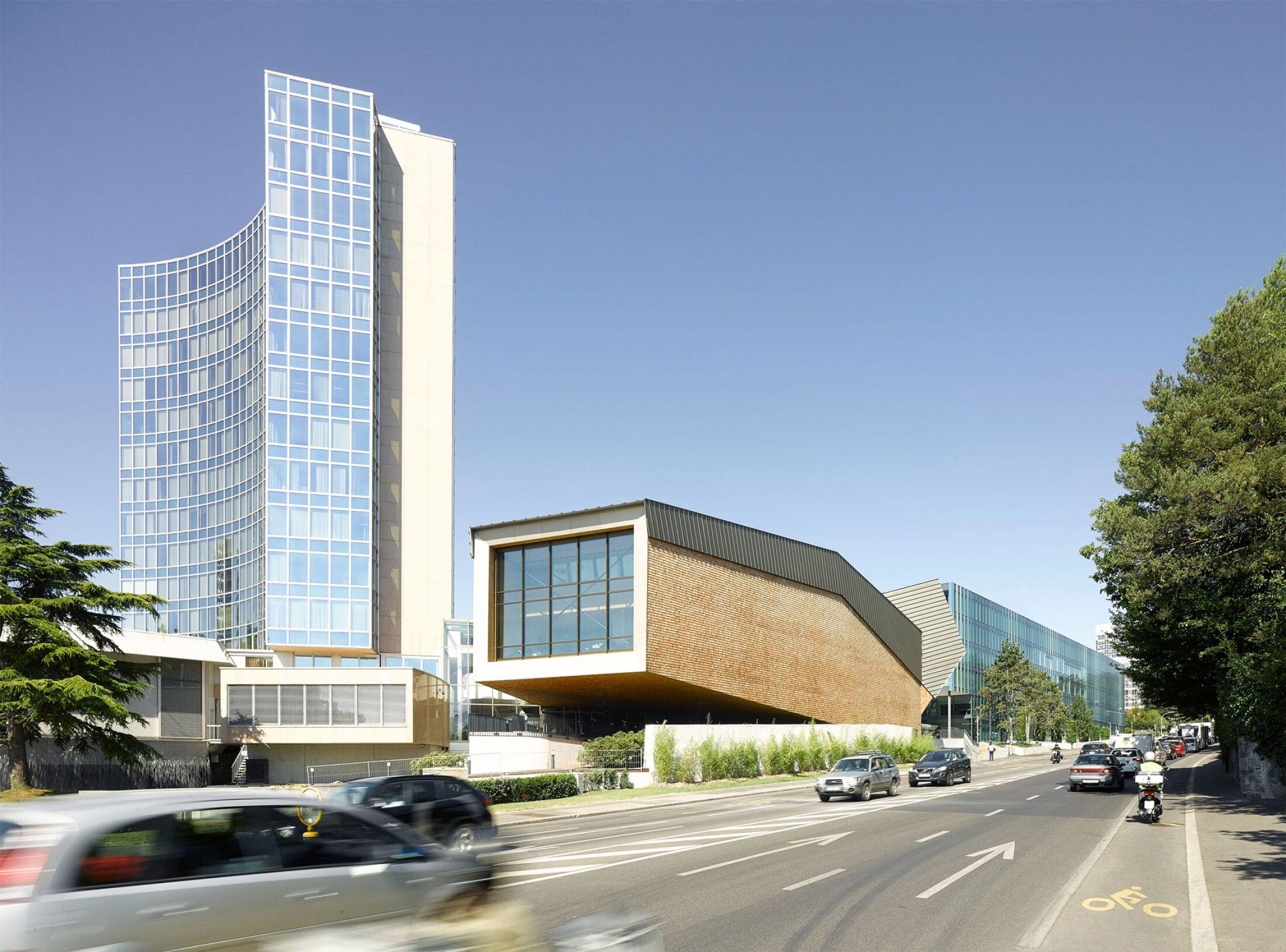 Project by Behnisch Architekturbuero, WIPO Conference Hall. Modern buildings along a street, including a curved glass tower and a cantilevered brick volume, with cars driving past.