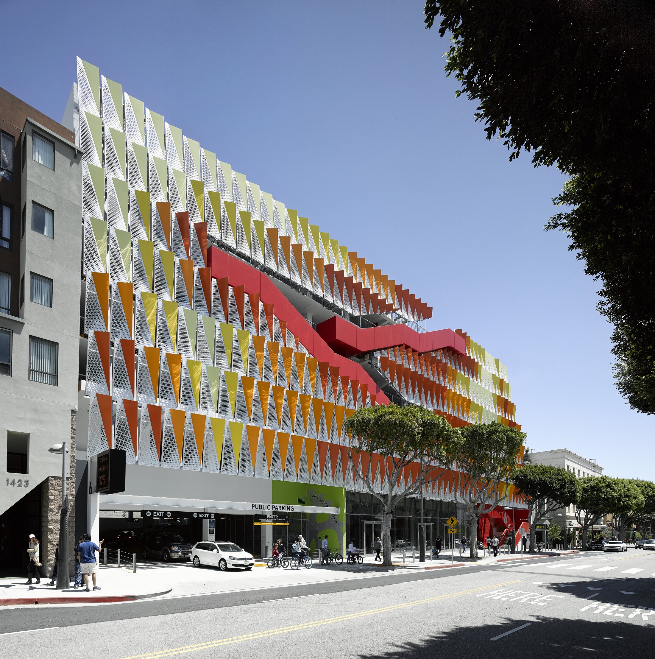 Project by Behnisch Architekturbuero, City of Santa Monica Parking Structure #6. Modern building with patterned facade and bold red exterior stair zigzagging across levels. Trees and pedestrians line the street in front. The facade is recessed around the red diagonal stair.