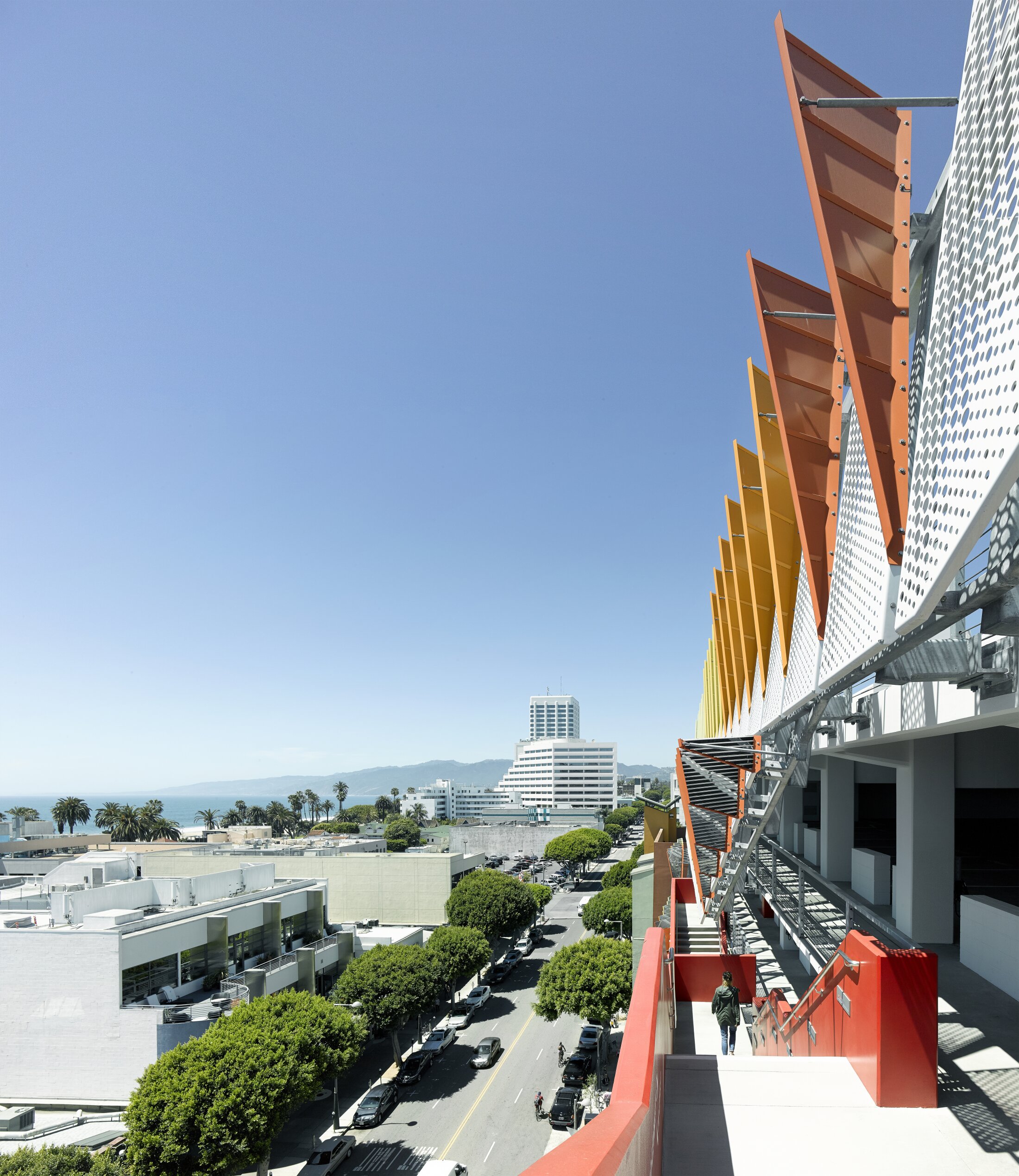 Project by Behnisch Architekturbuero, City of Santa Monica Parking Structure #6. Exterior walkway with red railing on a modern building, colorful vertical fins above, overlooking a street with trees and low-rise buildings.