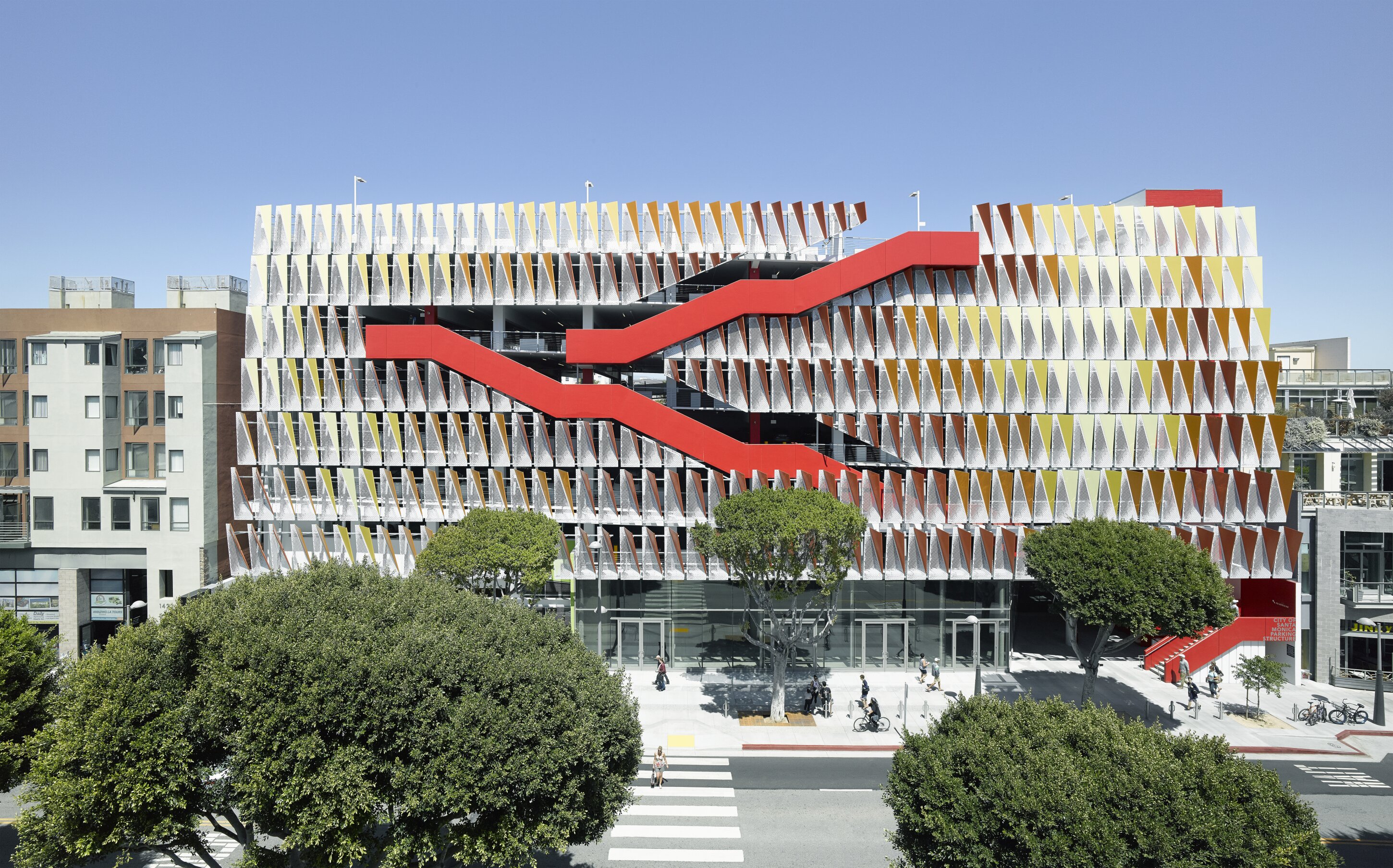 Project by Behnisch Architekturbuero, City of Santa Monica Parking Structure #6. Front view of the building. The patterned facade and the striking red exterior staircase, which zigzags across the levels, are clearly visible. In the foreground, trees and pedestrians crossing a zebra crossing can be seen.