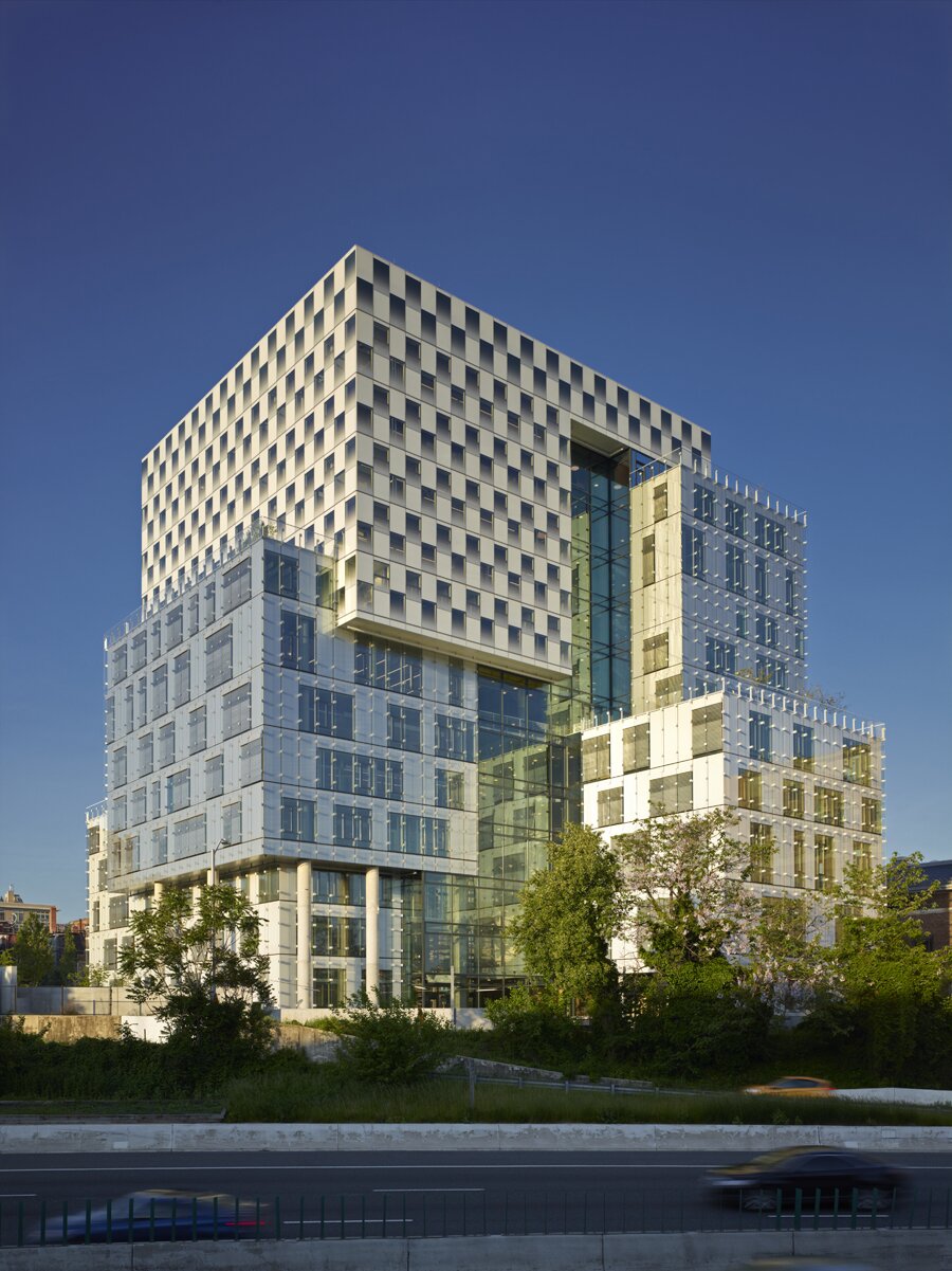 Project by Behnisch Architekturbuero, The John and Frances Angelos Law Center. Modern white office building surrounded by roads and trees. The exterior cladding reinforces the three-volume composition of the building through the use of three basic facade systems. For the office and classroom spaces, operable punched openings. The library facade features an operable curtain wall with a graduated frit pattern. The atrium facade is a multi-story glass wall attached to a steel frame.