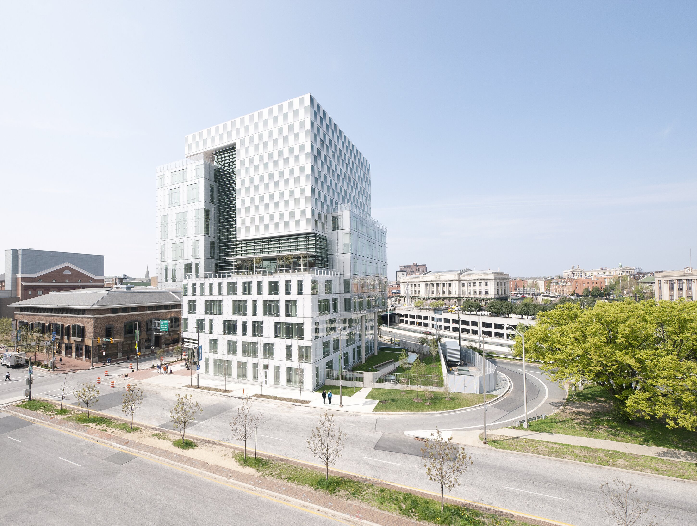 Project by Behnisch Architekturbuero, The John and Frances Angelos Law Center. Modern white office building surrounded by roads, trees, and nearby low-rise buildings. The exterior cladding reinforces the three-volume composition of the building through the use of three basic facade systems. For the office and classroom spaces, operable punched openings. The library facade features an operable curtain wall with a graduated frit pattern. The atrium facade is a multi-story glass wall attached to a steel frame