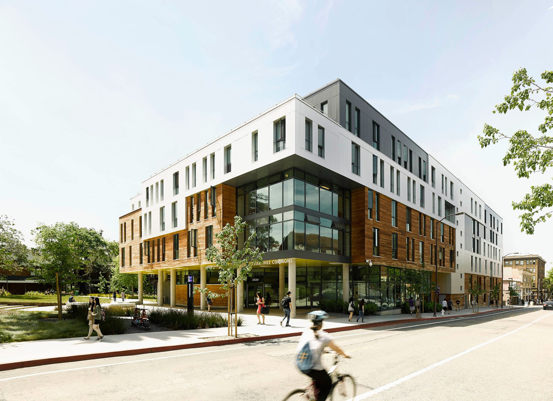 Project by Behnisch Architekturbuero, Maximino Martinez Commons, University of California. Modern multi-story building with white and wood facade next to a street. Pedestrians walk on sidewalks and a cyclist rides past in the foreground.