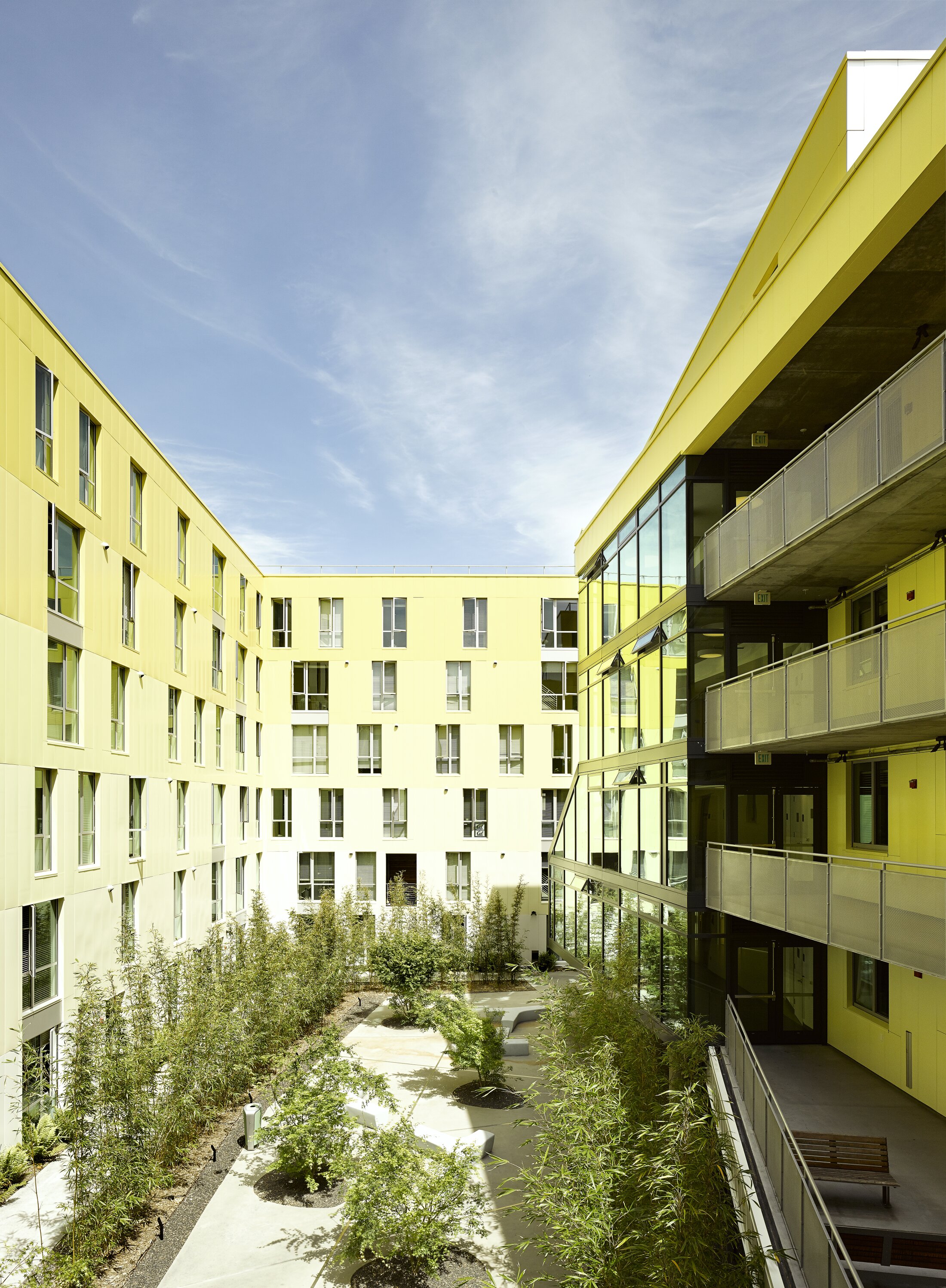 Project by Behnisch Architekturbuero, Maximino Martinez Commons, University of California. An enclosed courtyard between yellow buildings with balconies and glass facades. The courtyard is landscaped with trees and pathways and offers seating for residents.