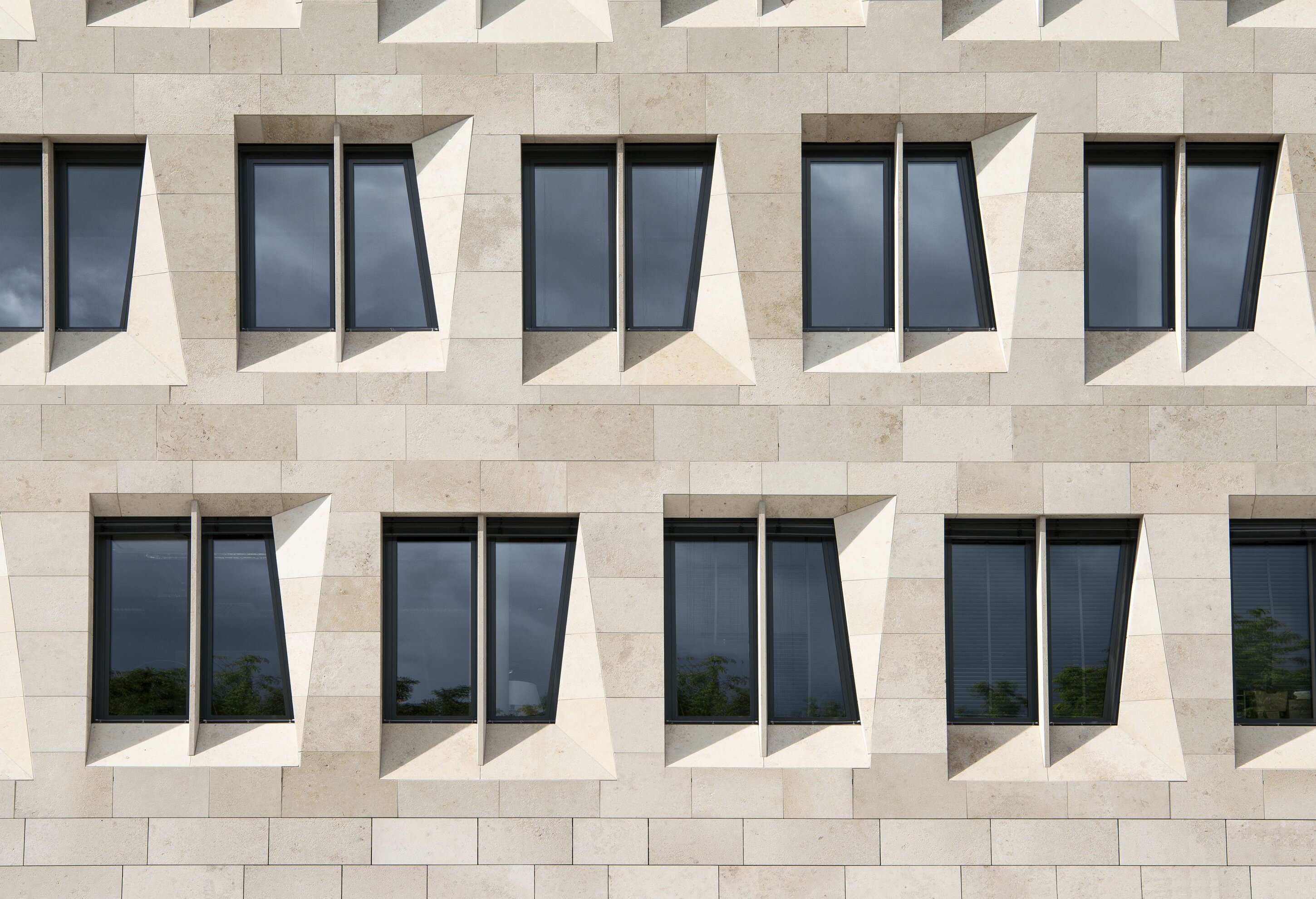 Project by Behnisch Architekturbuero, Dorotheen Quartier. Facade with beige stone panels and a grid of recessed, angular windows casting sharp shadows.