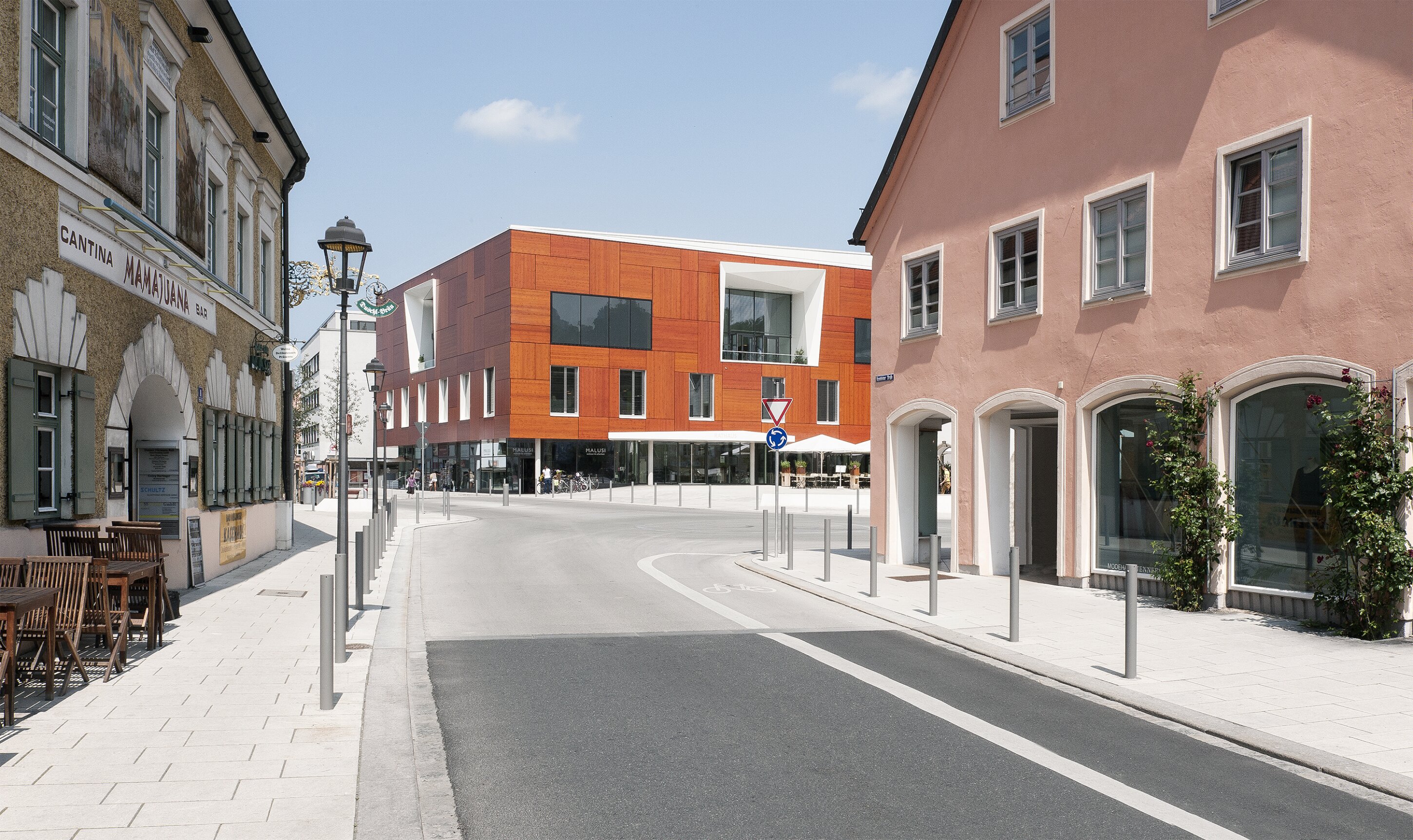 Project by Behnisch Architekturbuero, St. Benno School. Street view with older buildings and caf&eacute; seating, leading to a modern orange building at an intersection under a clear sky.