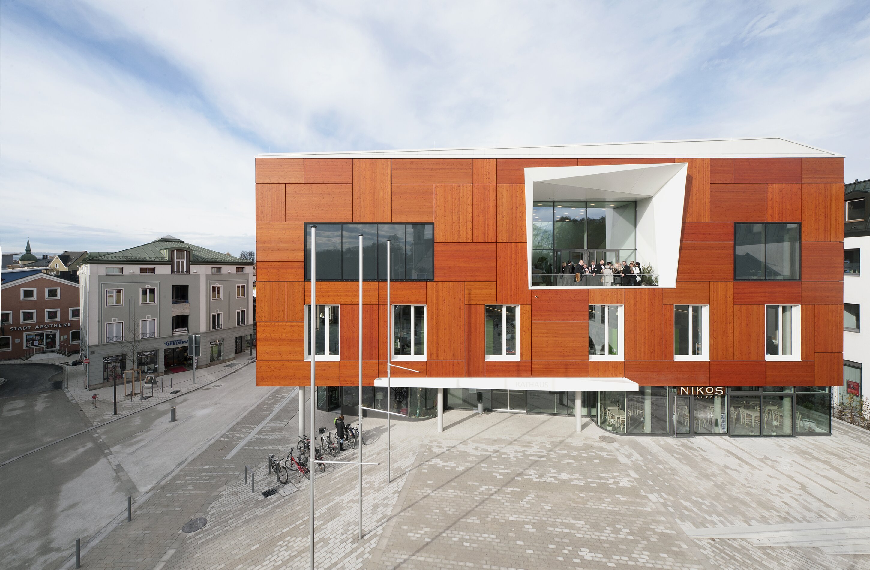 Project by Behnisch Architekturbuero, Townhall Bad Aibling. Modern red-clad building on a plaza with large windows and a recessed balcony, next to smaller older buildings along a quiet street.