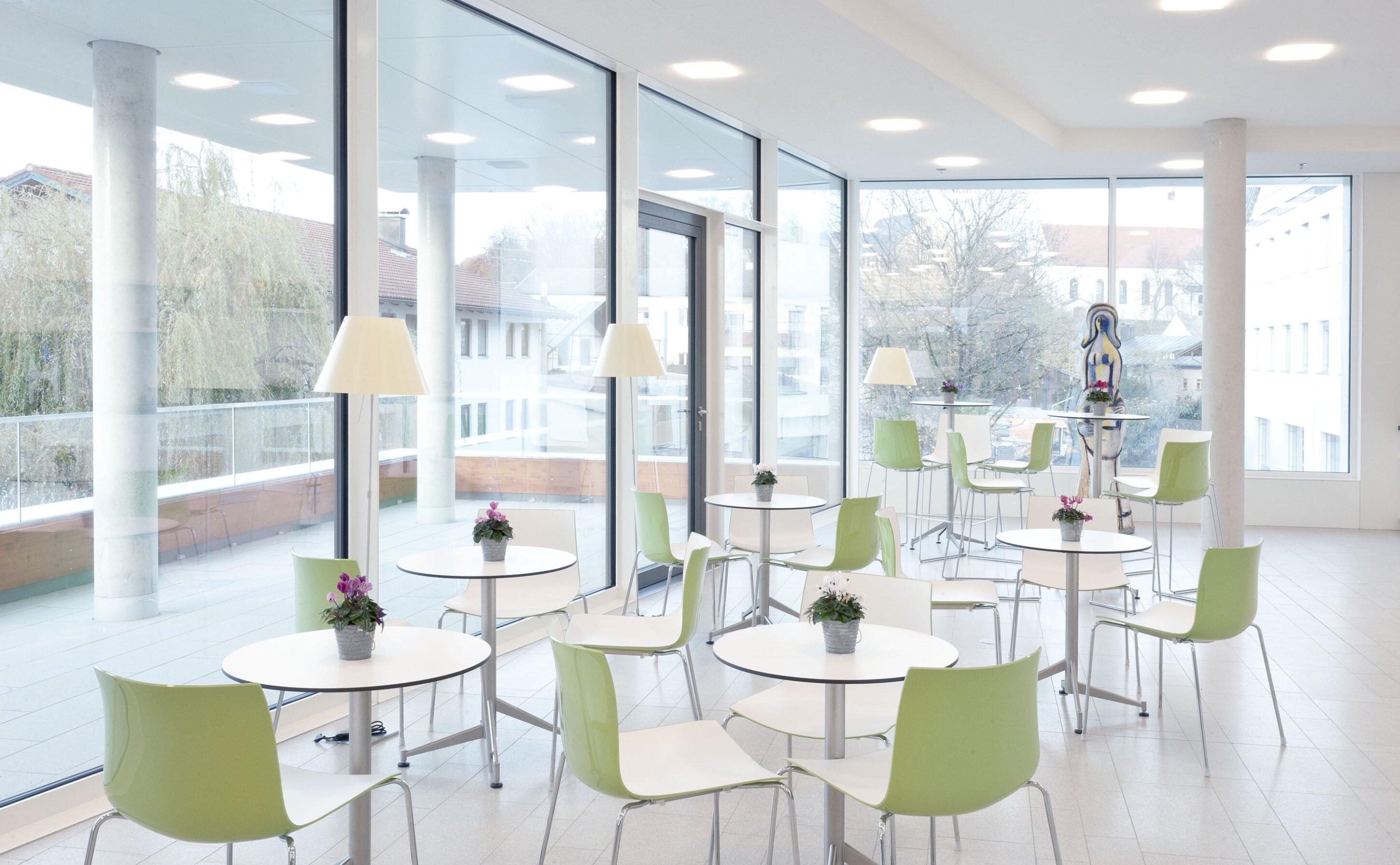 Project by Behnisch Architekturbuero, Townhall Bad Aibling. Bright lounge with round tables, green chairs, potted flowers, and floor-to-ceiling windows overlooking nearby buildings.