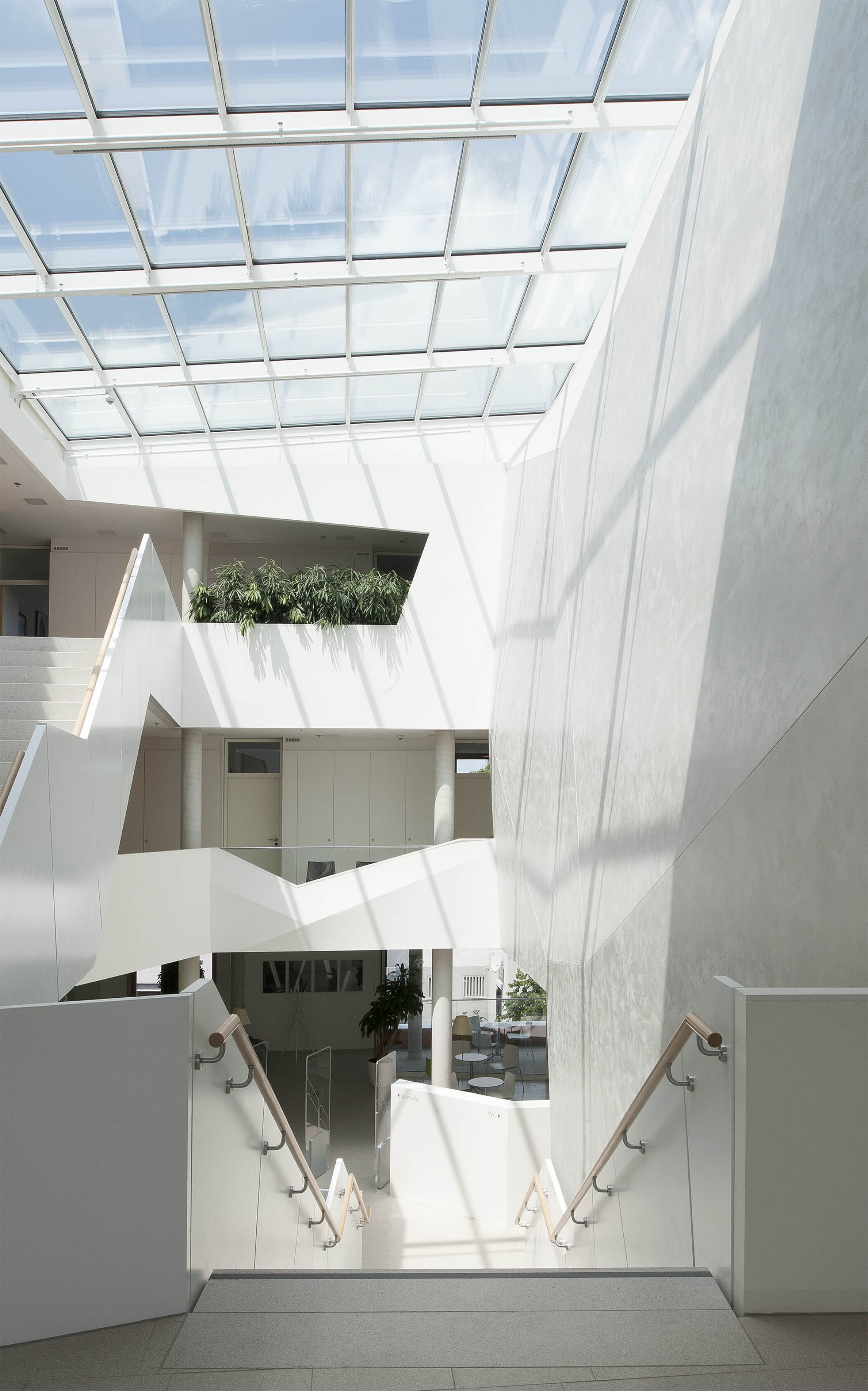 Project by Behnisch Architekturbuero, Townhall Bad Aibling. Bright white atrium with glass roof, staircases, and railings, sunlight casting shadows across walls and floors.