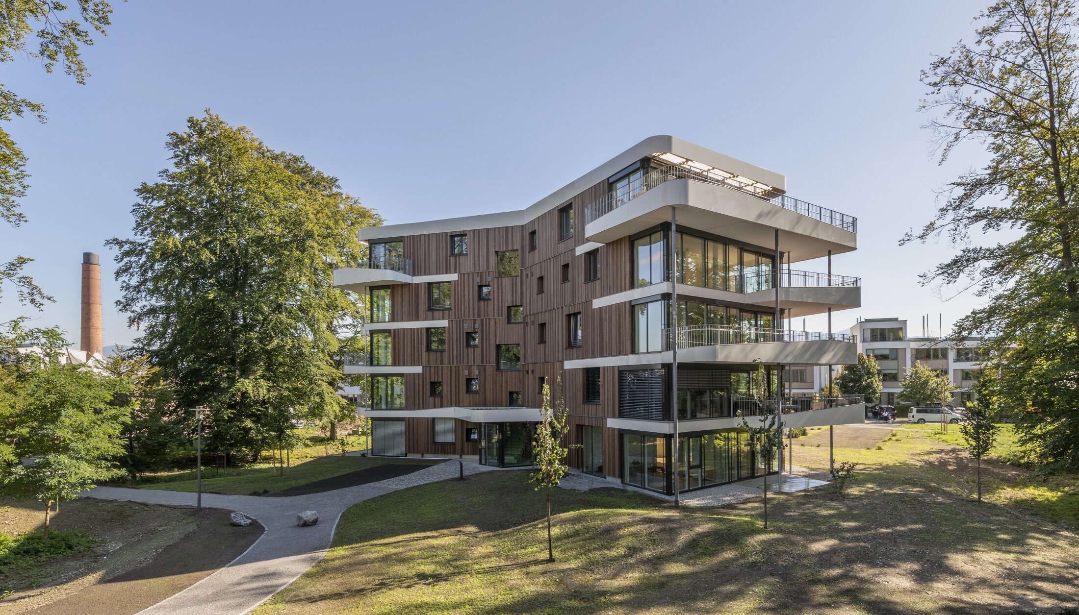 Project by Behnisch Architekturbuero, Y-Houses. A modern, multi-story residential building with a wooden facade and surrounding white balconies, set in a green park. A brick chimney is visible in the background.