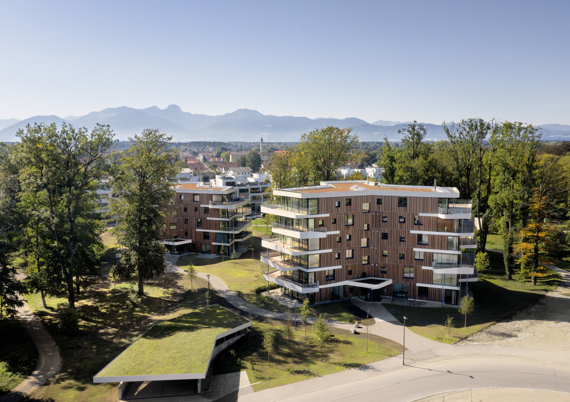 Project by Behnisch Architekturbuero, Y-Houses.Two modern apartment buildings with wooden facades and balconies, nestled in a well-maintained green area with a mountain panorama in the background.