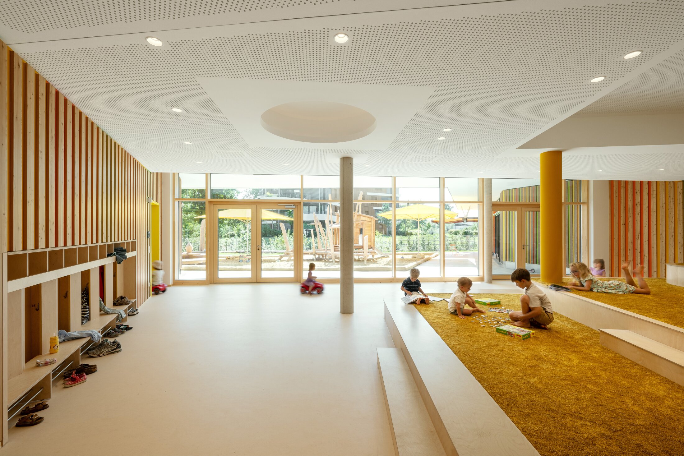 Project by Behnisch Architekturbuero, Conradty-Houses. Bright, welcoming daycare interiors with children playing on a raised carpeted area. A playground outside is visible through the large windows.