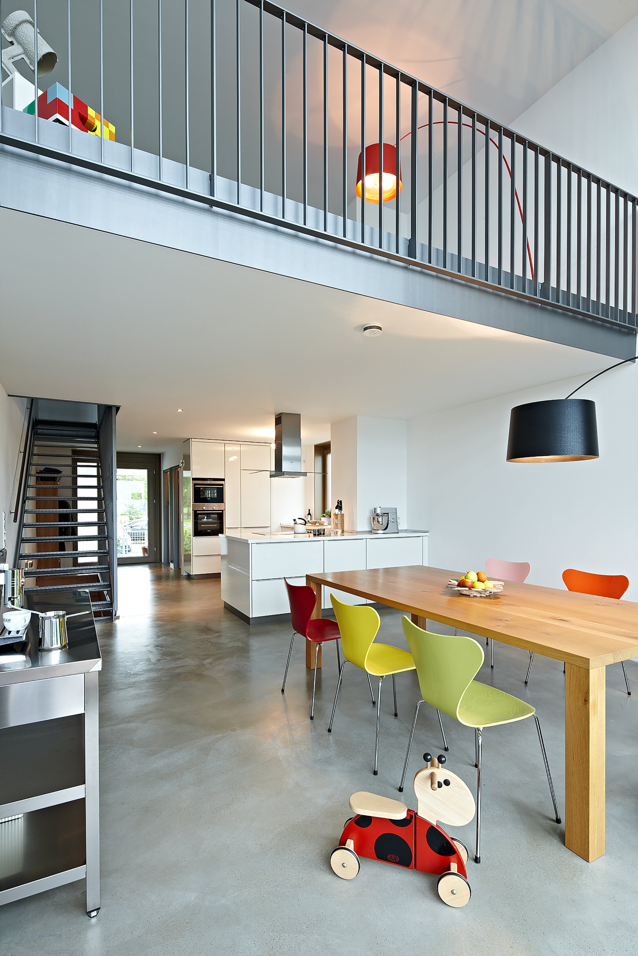 Project by Behnisch Architekturbuero, Loft Houses Kolbermoor. Modern open-plan kitchen and dining area with wooden table, colorful chairs, and mezzanine above. An impression into the interior of one of the Loft Houses.