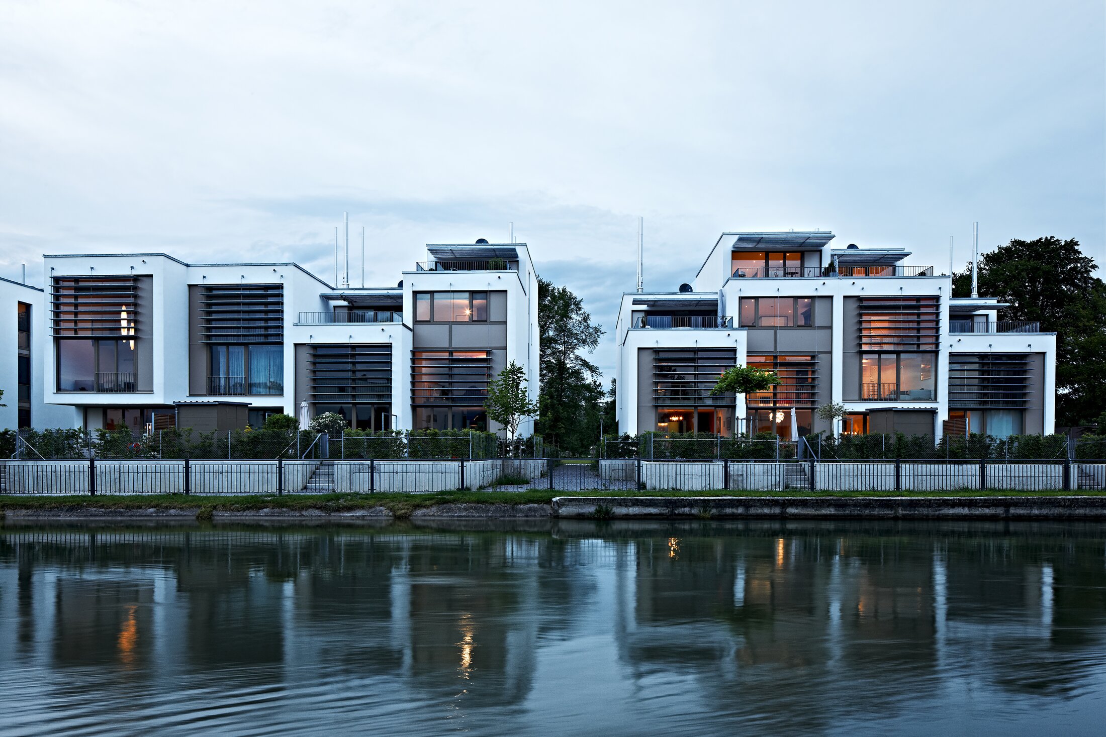 Project by Behnisch Architekturbuero, Loft Houses Kolbermoor. Front view of the modern white Loft Houses along the Mangfall Canal in an evening atmosphere.