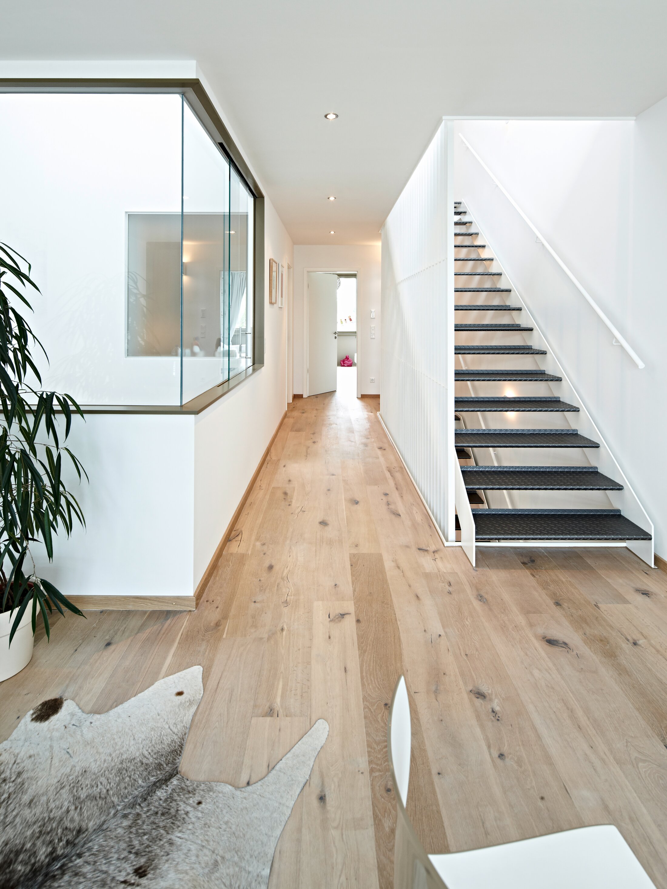 Project by Behnisch Architekturbuero, Loft Houses Kolbermoor. A bright, modern hallway with a wooden floor, a glass wall facing the courtyard, and an open staircase leading to the upper floor. An impression into the interior of one of the Loft Houses.
