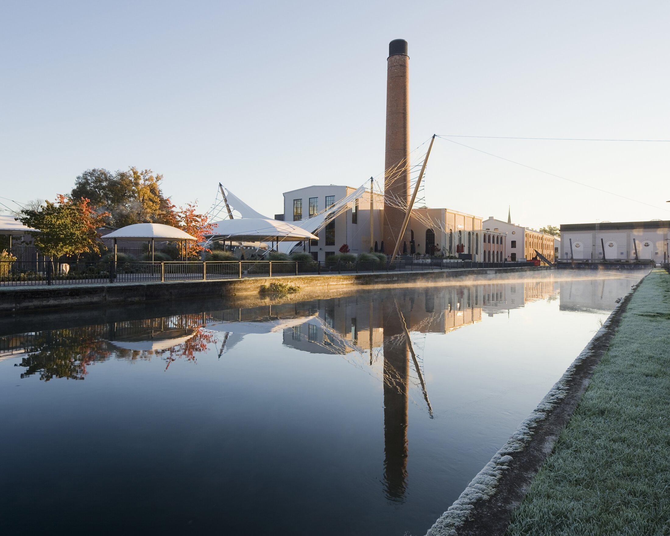 Project by Behnisch Architekturbuero, QuestForum and Rosengarten. QuestForum and rose garden with a brick chimney in the background. Flexible event space covered by a rope net structure. Everything is reflected in the tranquil canal.