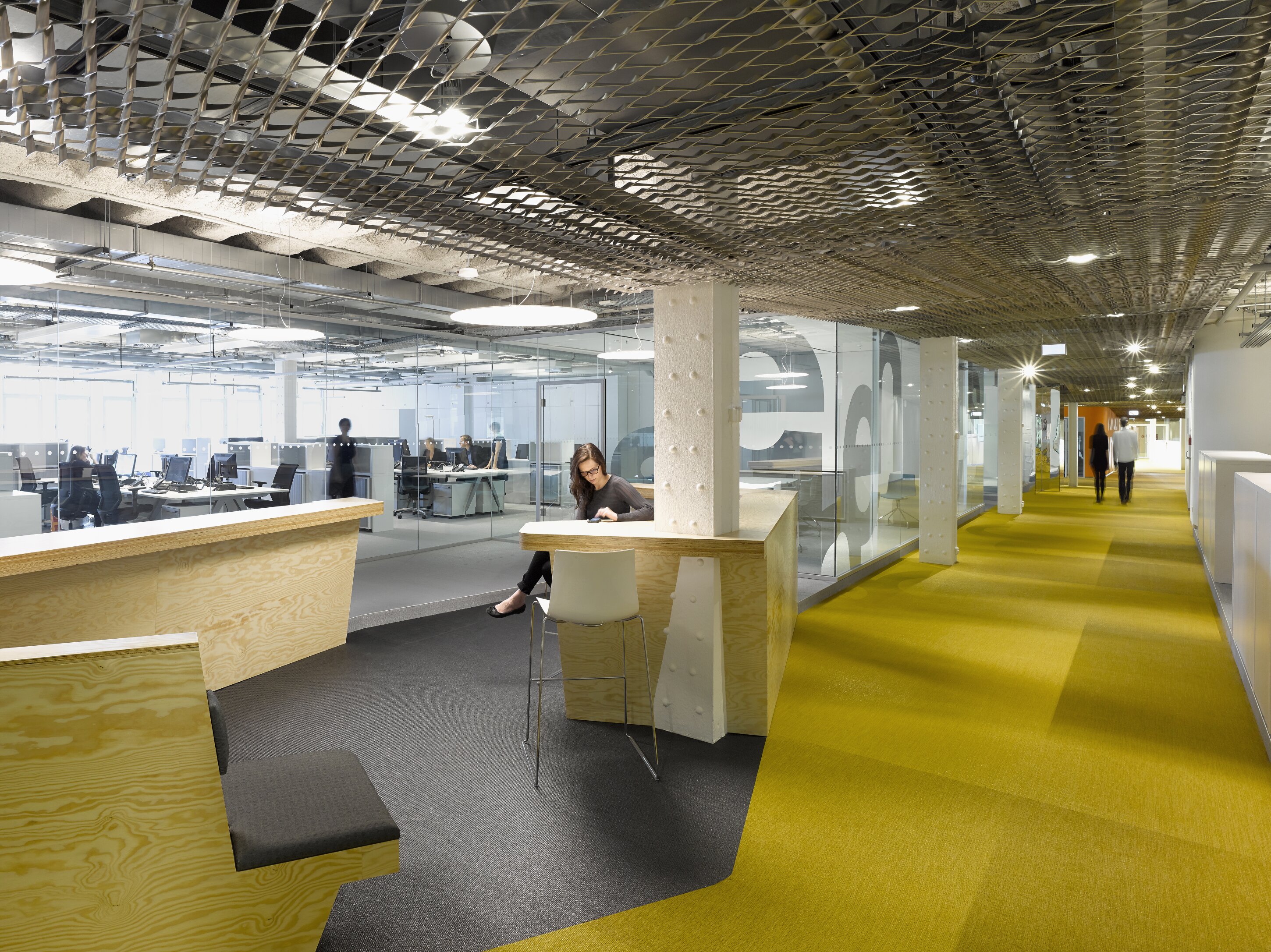Project by Behnisch Architekturbuero, Breuninger Headquarter. Open office corridor with yellow carpet, mesh ceiling, glass-walled work area, and people working and walking; woman seated at a wooden counter.