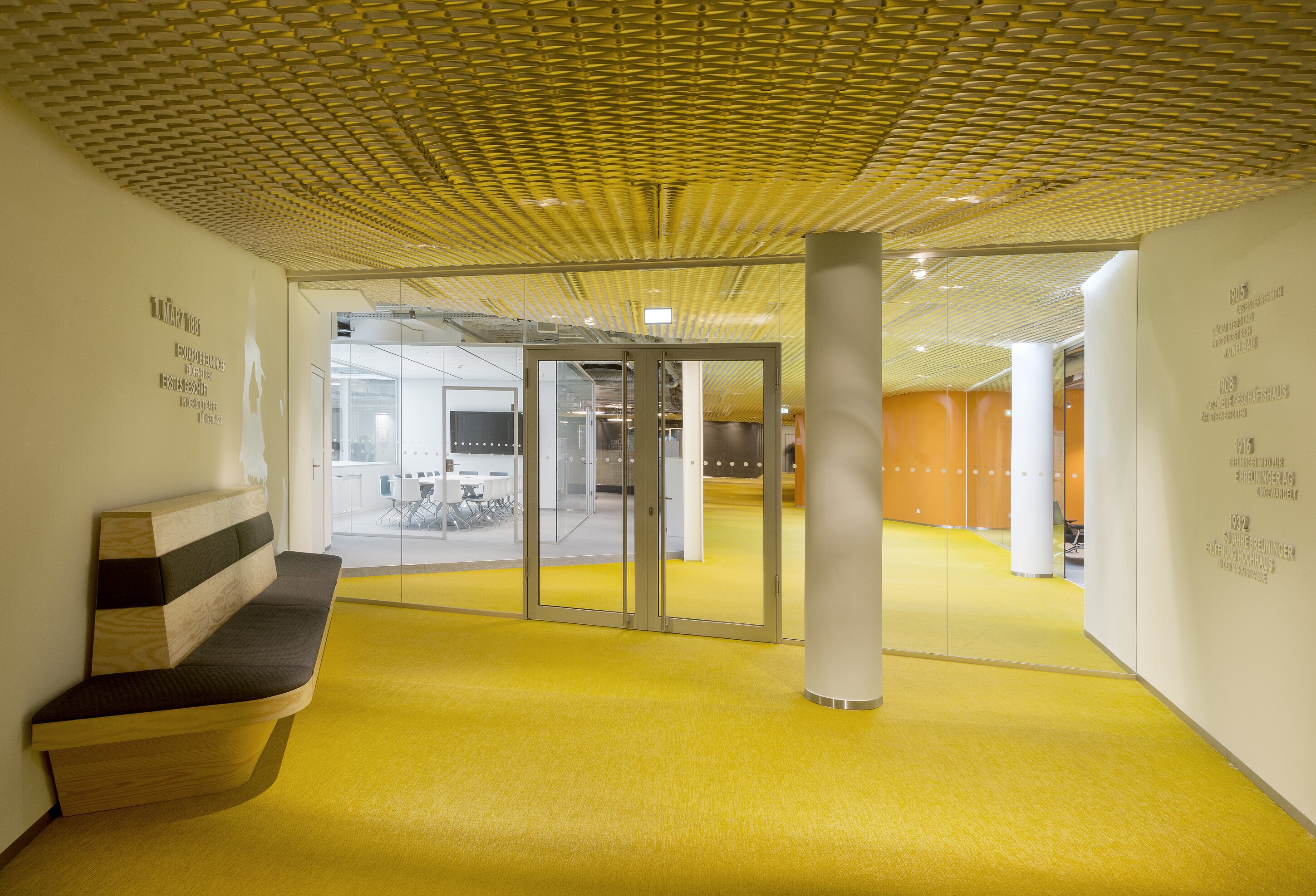 Project by Behnisch Architekturbuero, Breuninger Headquarter. Interior hallway with bright yellow floor and textured ceiling, glass doors, a bench on the left, and meeting rooms visible beyond.