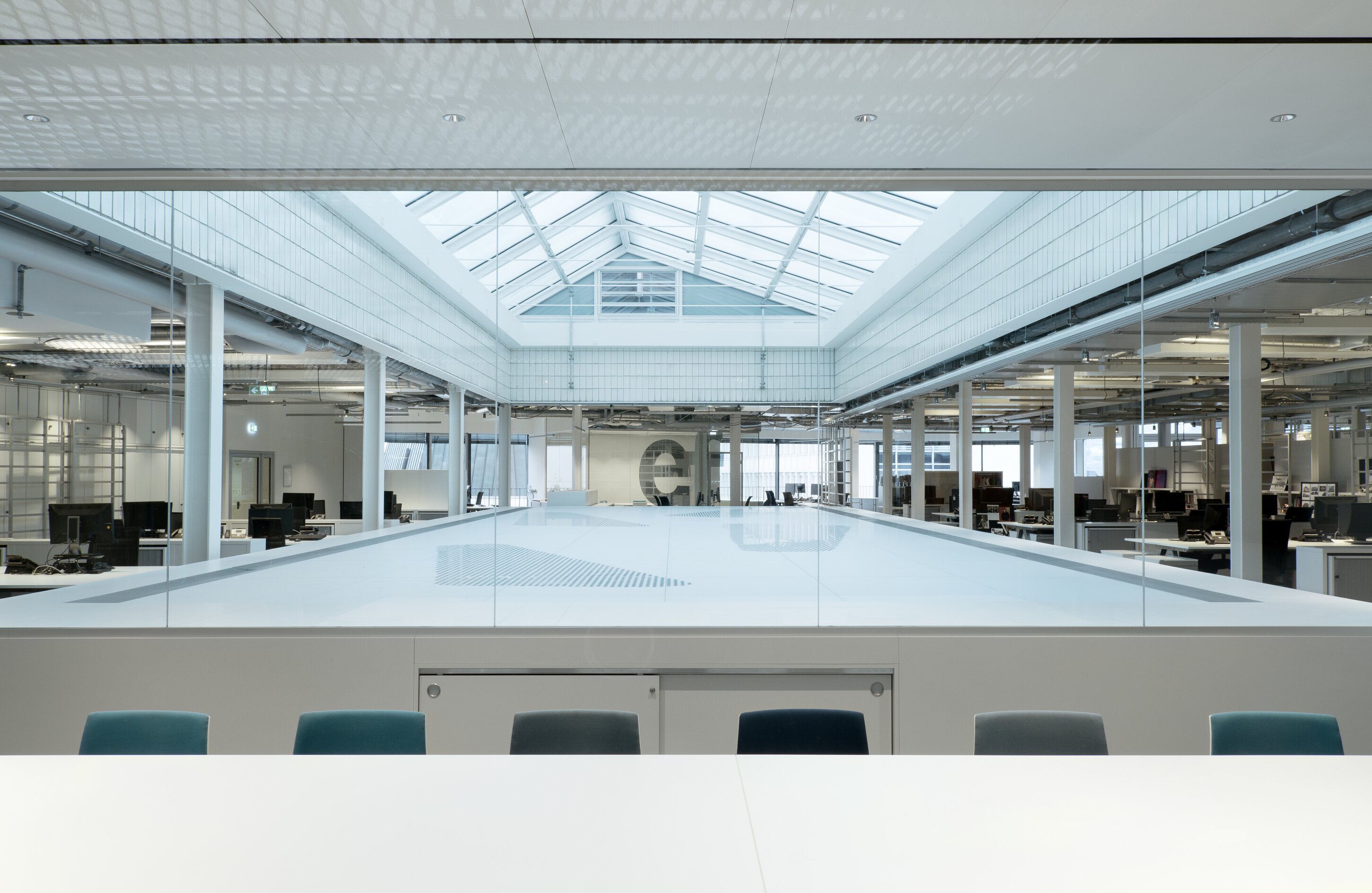 Project by Behnisch Architekturbuero, Breuninger Headquarter. Open-plan office interior with skylight, glass railing, desks and computers around a central lightwell, viewed from a meeting table with chairs in foreground.
