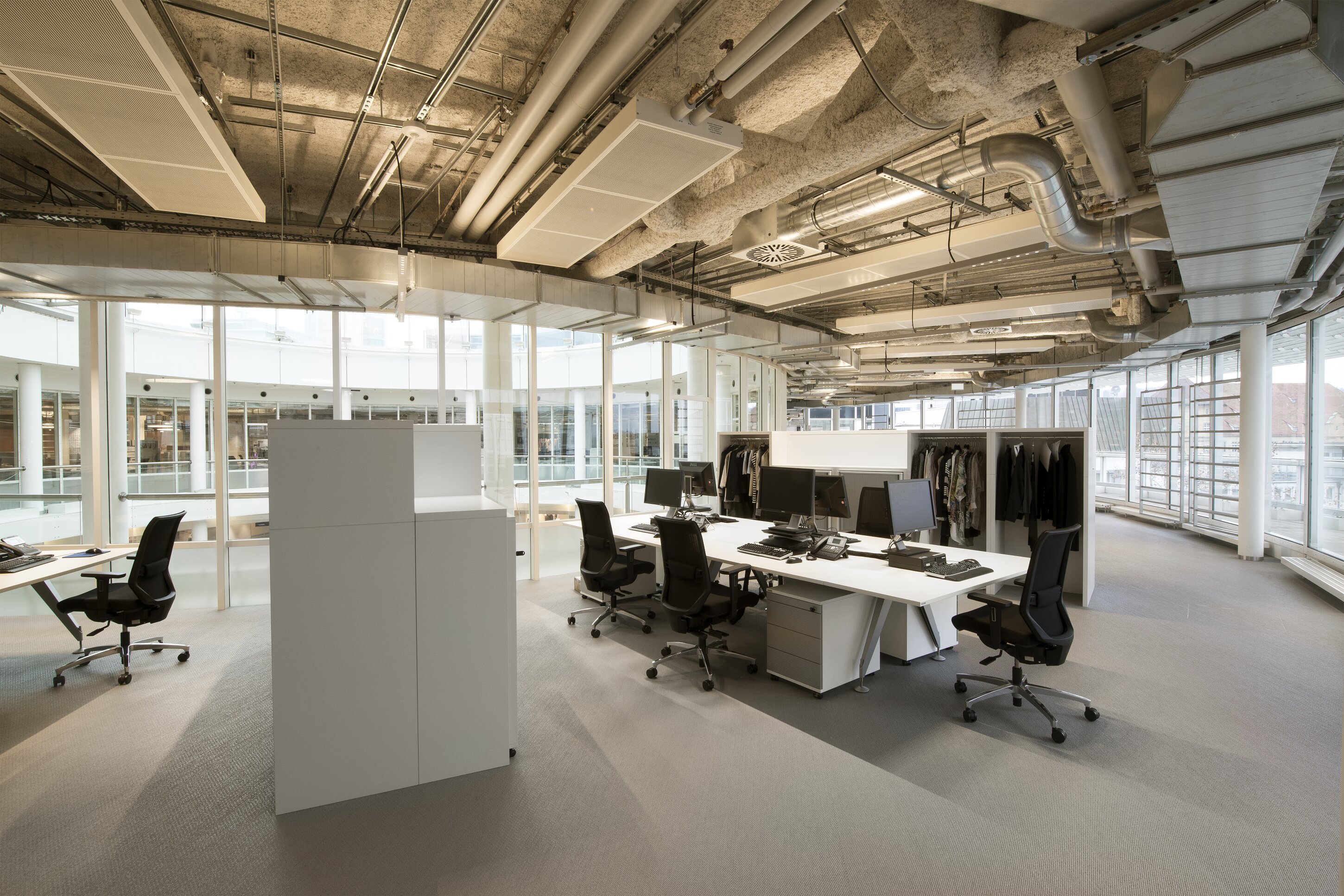 Project by Behnisch Architekturbuero, Breuninger Headquarter. Open-plan office with desks, computers, office chairs, clothing racks, and exposed ceiling pipes beside large windows.
