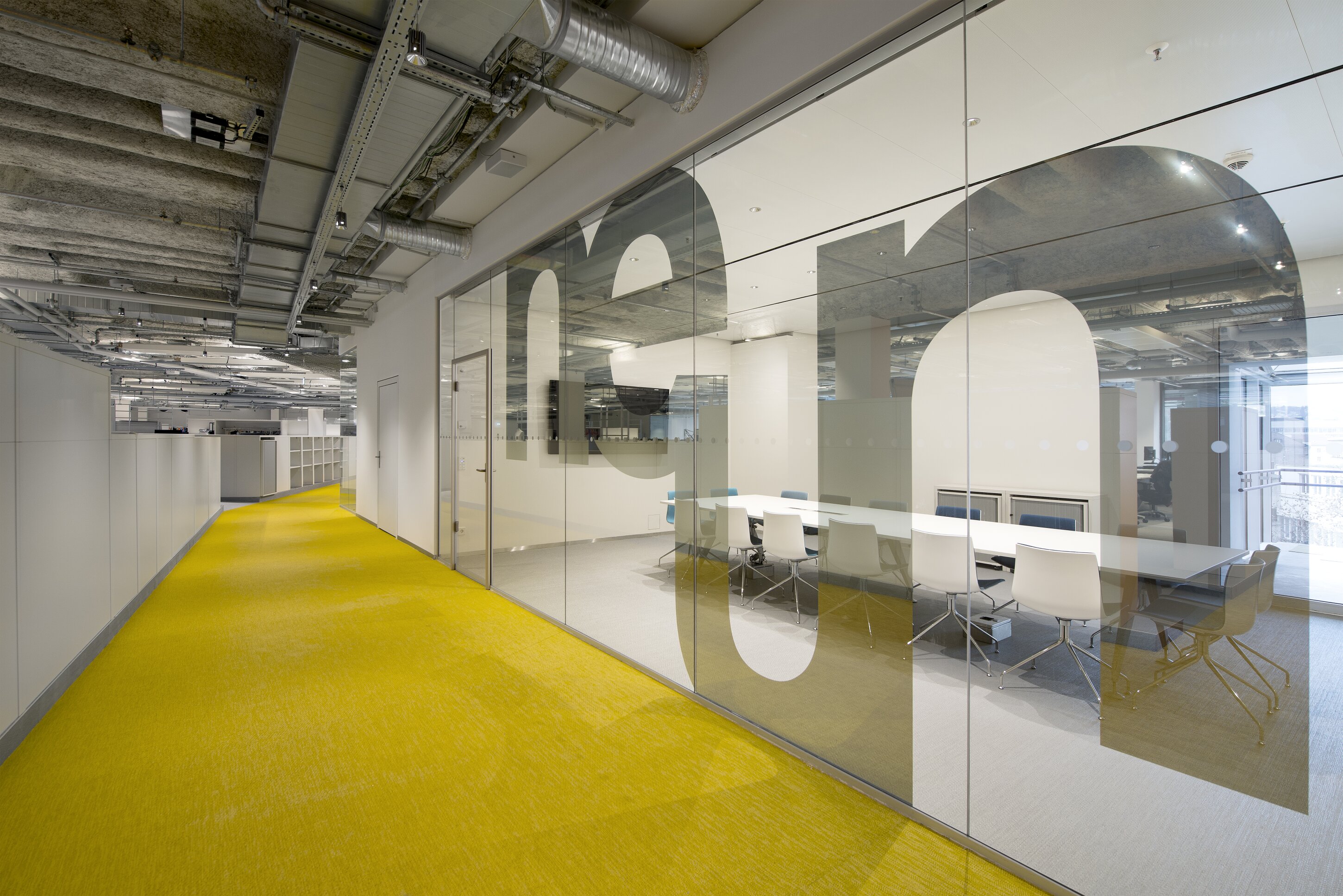 Project by Behnisch Architekturbuero, Breuninger Headquarter. Office corridor with yellow carpet, exposed ceiling, and glass-walled meeting room with long table and white chairs.