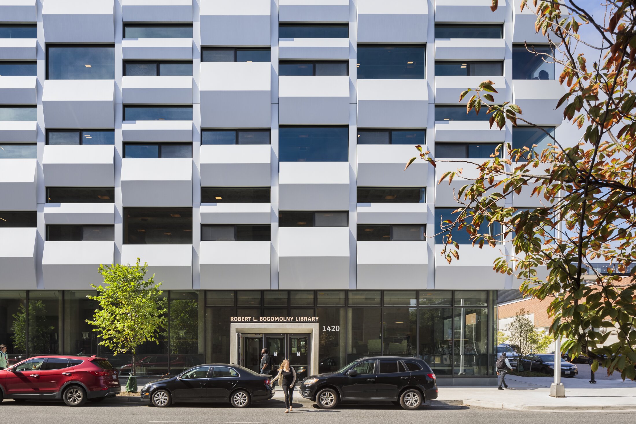 Project by Behnisch Architekturbuero, Robert Bogomolny Library. Modern library facade with patterned white panels, street-level entrance, parked cars, and pedestrians on sidewalk under trees.