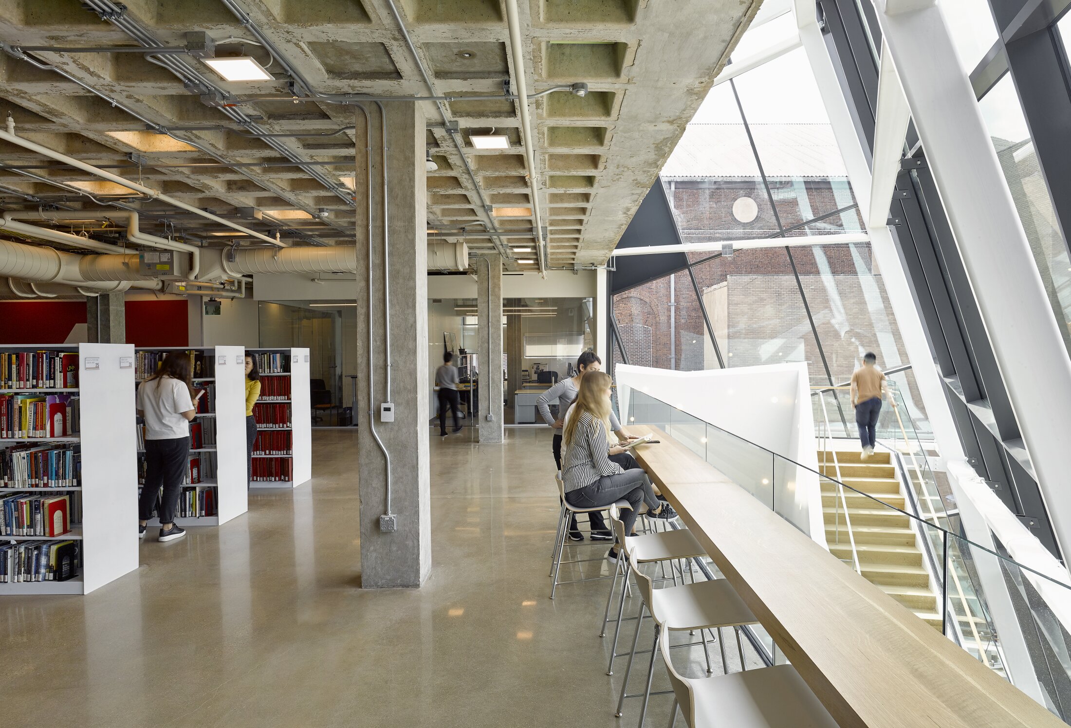 Project by Behnisch Architekturbuero, Robert Bogomolny Library. Library interior with bookshelves, exposed concrete ceiling, and window seating for reading. Behind it, a staircase and glass facade allow light to flood the room.