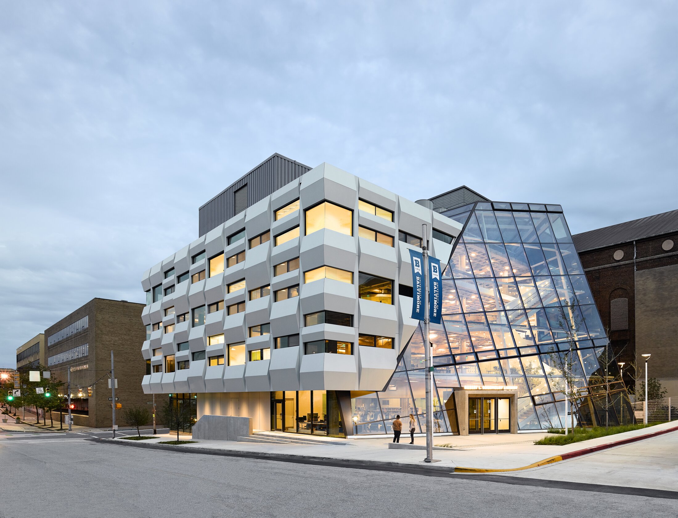 Project by Behnisch Architekturbuero, Robert Bogomolny Library. Modern multi-story building with a faceted white facade and glass atrium on a street corner. The interior lights glowing at dusk.
